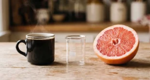 Un pomelo rosa cortado por la mitad junto a una taza de café negro humeante sobre una mesa de madera clara.
